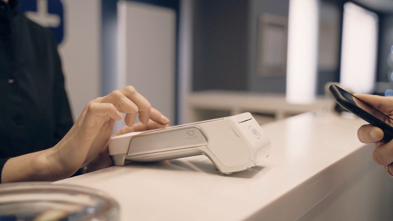 Hand using a white ShapesPay terminal at a bright checkout counter